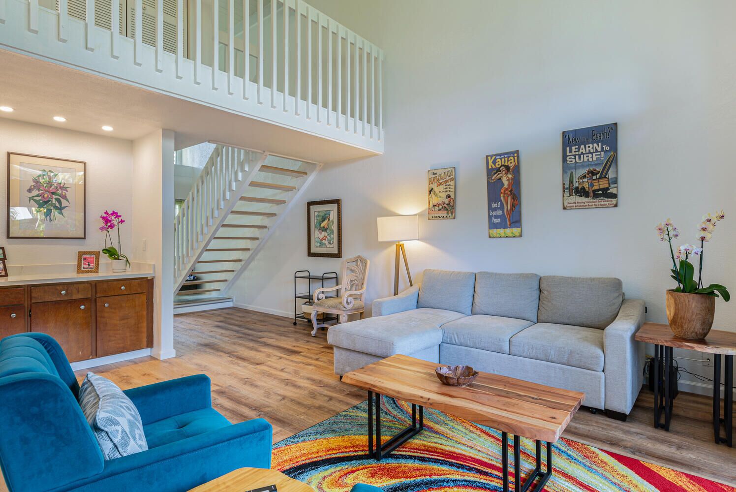 Ein geräumiges Wohnzimmer mit Treppe, einem blauen Sessel, einem grauen Sofa und einem bunten Teppich.