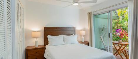 Un dormitorio con cabecera de madera, cama con ropa blanca, dos lámparas de noche, ventilador de techo y puerta corrediza de vidrio que da a un balcón con plantas.