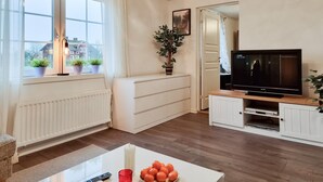 Een woonkamer met een witte radiator, een televisie op een houten dressoir, een plant en een raam met gordijnen.