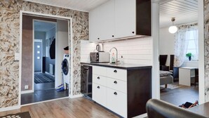 Een moderne keuken met witte kasten, een ingebouwde oven en een koffiezetapparaat. Er ligt een houten vloer en er is uitzicht op een zithoek met een bank en een raam.