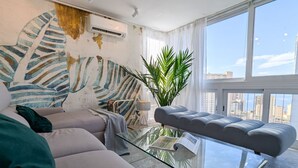 Ein modernes Wohnzimmer mit einem Glastisch, einem grauen Sofa, einer großen Pflanze und Blick auf die Stadt durch große Fenster.