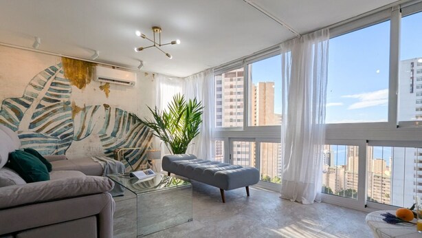 Ein modernes Wohnzimmer mit einer Couch, einem grauen Hocker, einem Glastisch und einem großen Fenster mit Vorhängen.