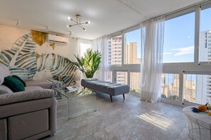 Een moderne woonkamer met een bank, een grijze poef, een glazen salontafel en een groot raam met gordijnen.
