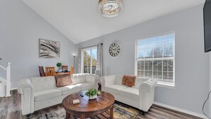A spacious living room with a white sofa, a round wooden coffee table, and a large window with blinds. - Home of Light, Branson, Mo (Branson)