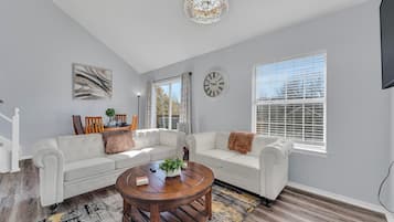 A spacious living room with a white sofa, a round wooden coffee table, and a large window with blinds.