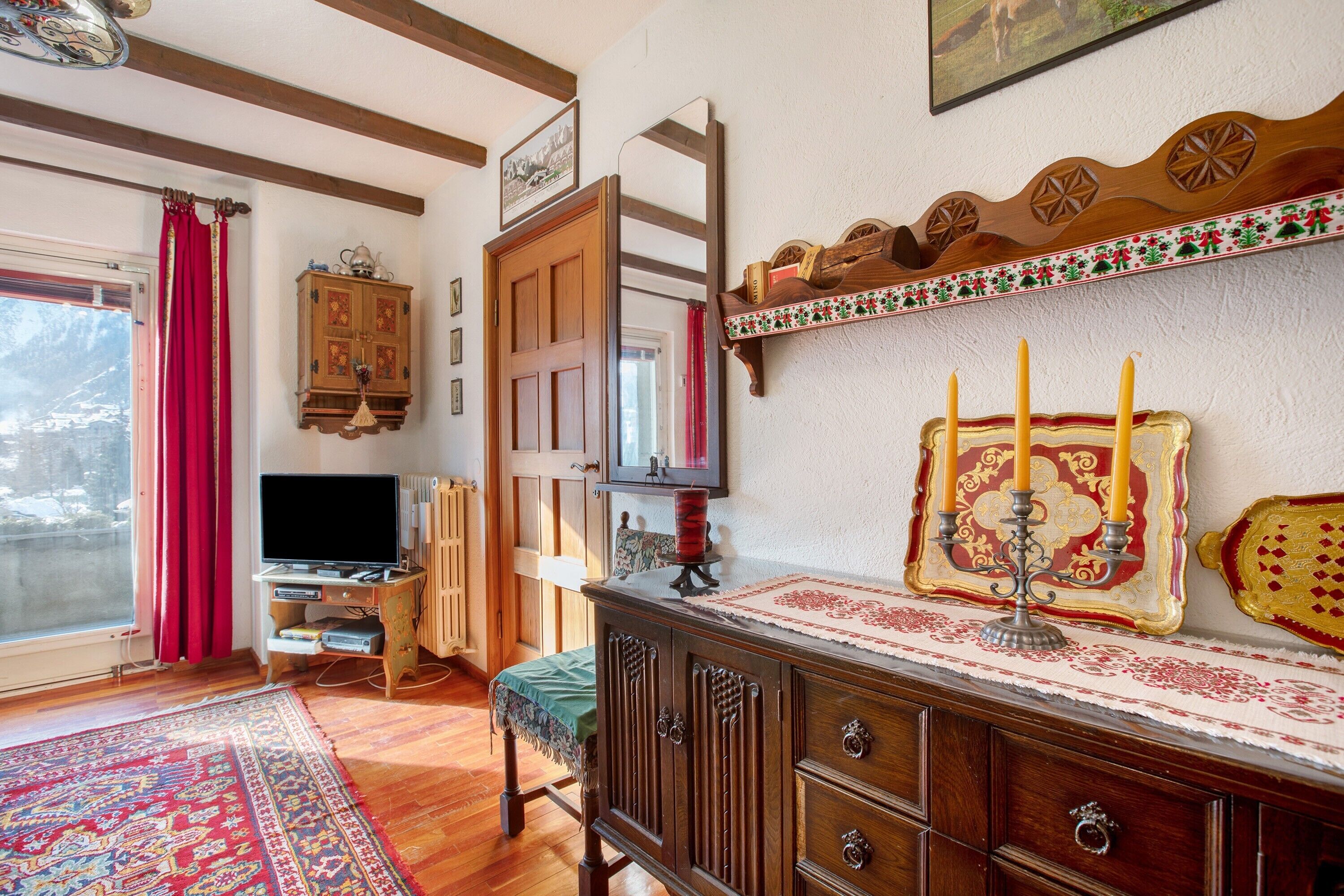 Habitació amb un armari de fusta, televisió, cortina vermella, alfàbregua estampada i una estantanya penjada a la paret amb un objecte decoratiu.