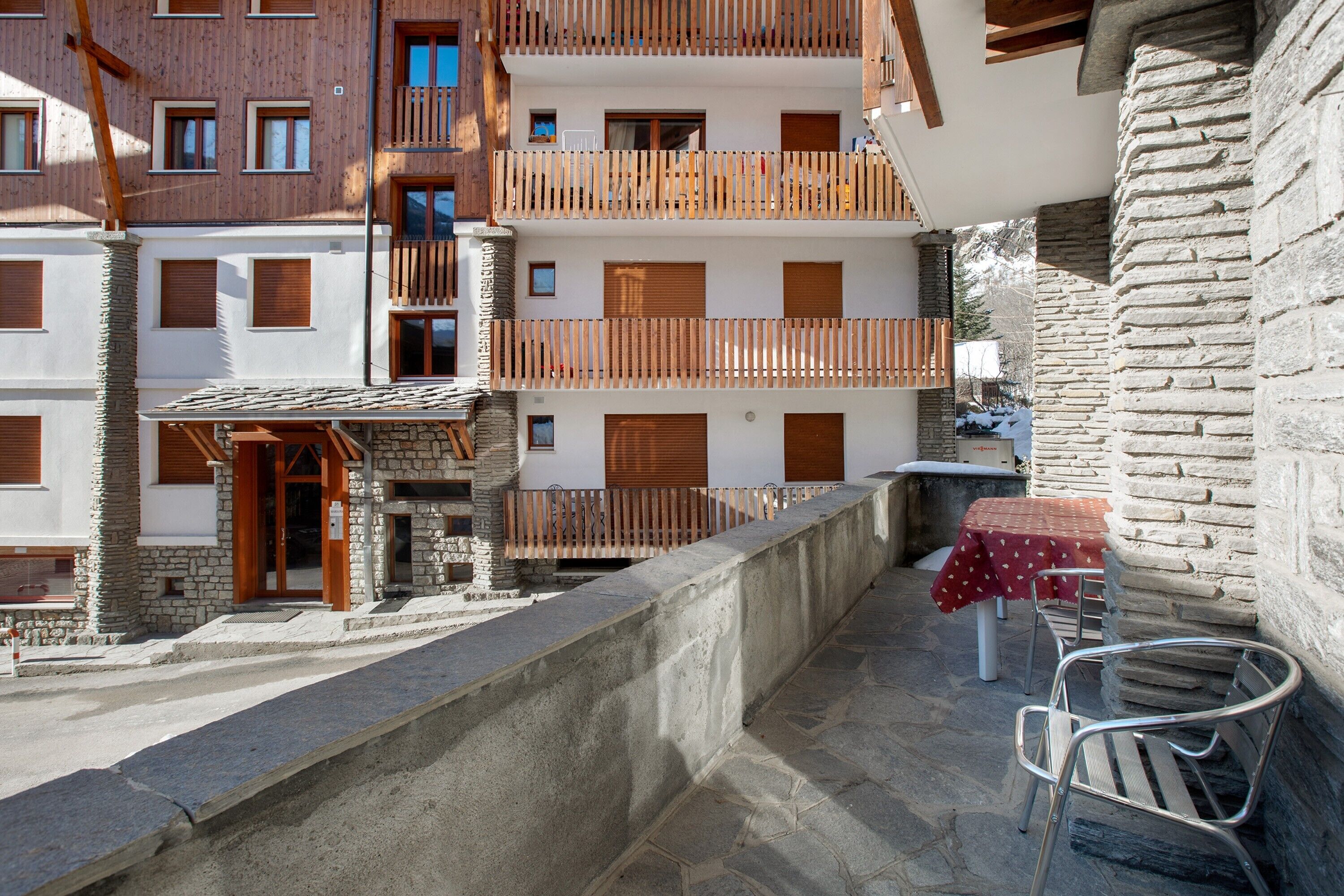 A multi-story building with balconies, stone walls, and a table with chairs on the ground floor.