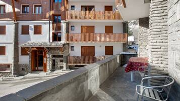 A multi-story building with balconies, stone walls, and a table with chairs on the ground floor.