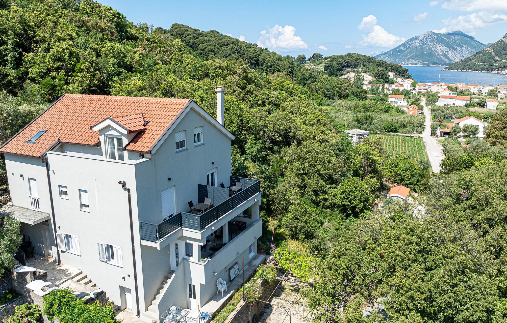 Un bâtiment à deux étages, doté d’un toit recouvert de tuiles rouges, d’un balcon et offrant une vue sur une zone côtière avec des maisons et une étendue d’eau.