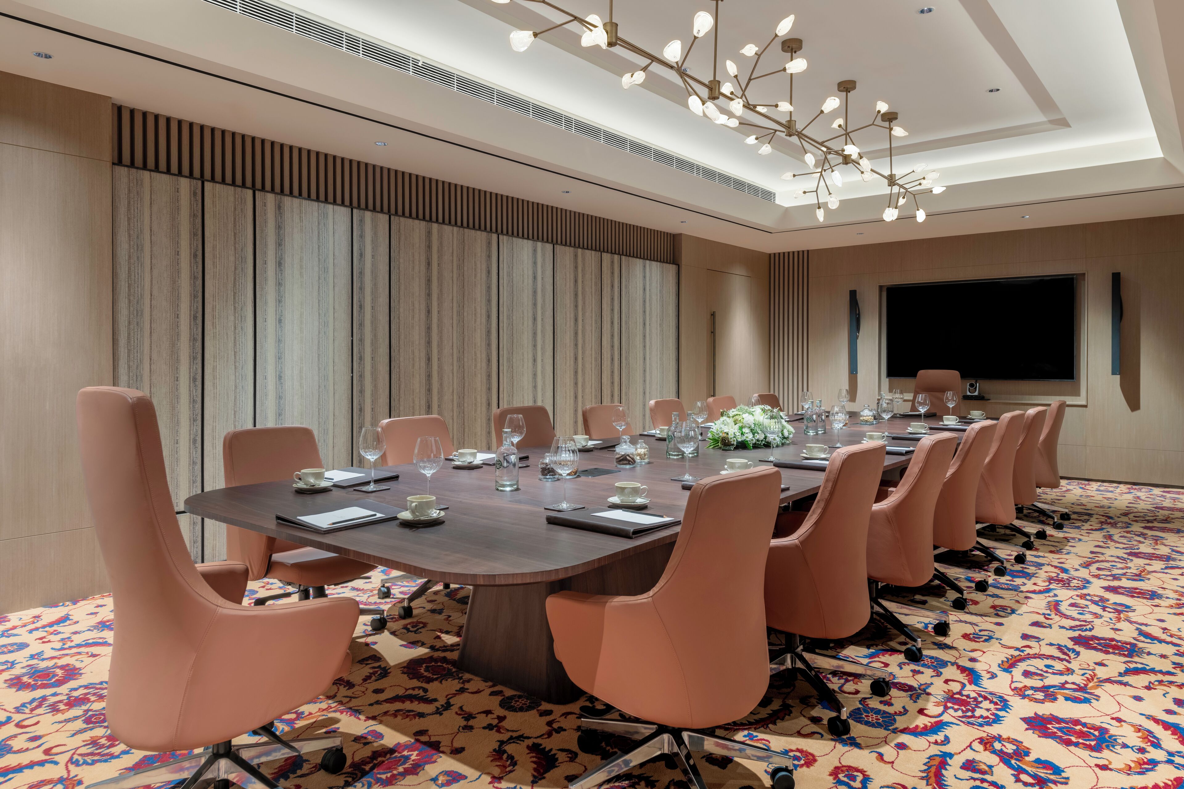A conference room with a long table, multiple chairs, a large screen, and a patterned carpet.