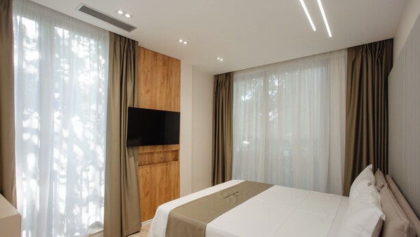 Une chambre d’hôtel moderne dotée d’un grand lit, d’un téléviseur à écran plat fixé au mur et de grandes fenêtres avec des rideaux légers.