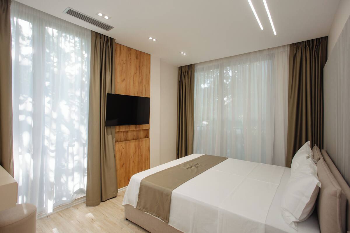 A modern hotel room with a large bed, a flat-screen TV mounted on the wall, and large windows with sheer curtains.