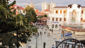 Une terrasse extérieure avec deux chaises à armature métallique noire, une table en verre et une vue sur une rue de la ville, avec des immeubles et un bâtiment à toit rouge en arrière-plan.