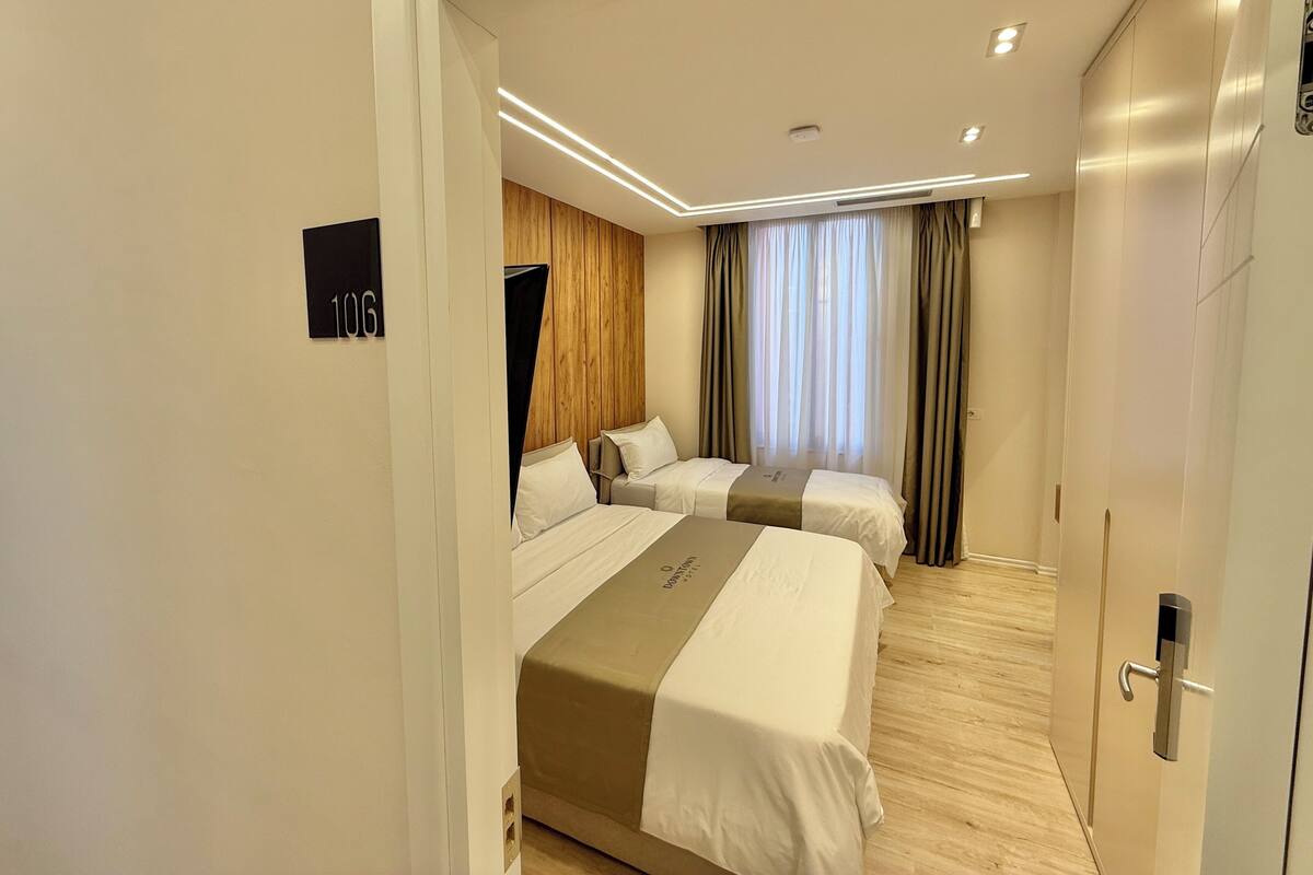 A hotel room with two beds, a wooden accent wall, a large window with curtains, and a door with a modern handle.