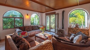 A cozy living room with a sofa, armchairs, a coffee table, and a fireplace. Large windows offer views of greenery outside. - Casa Surrender Cómpeta by Ruralidays (Málaga)