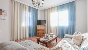 Un salon avec un canapé, une table basse en bois, un téléviseur posé sur un meuble en bois et des rideaux bleus.