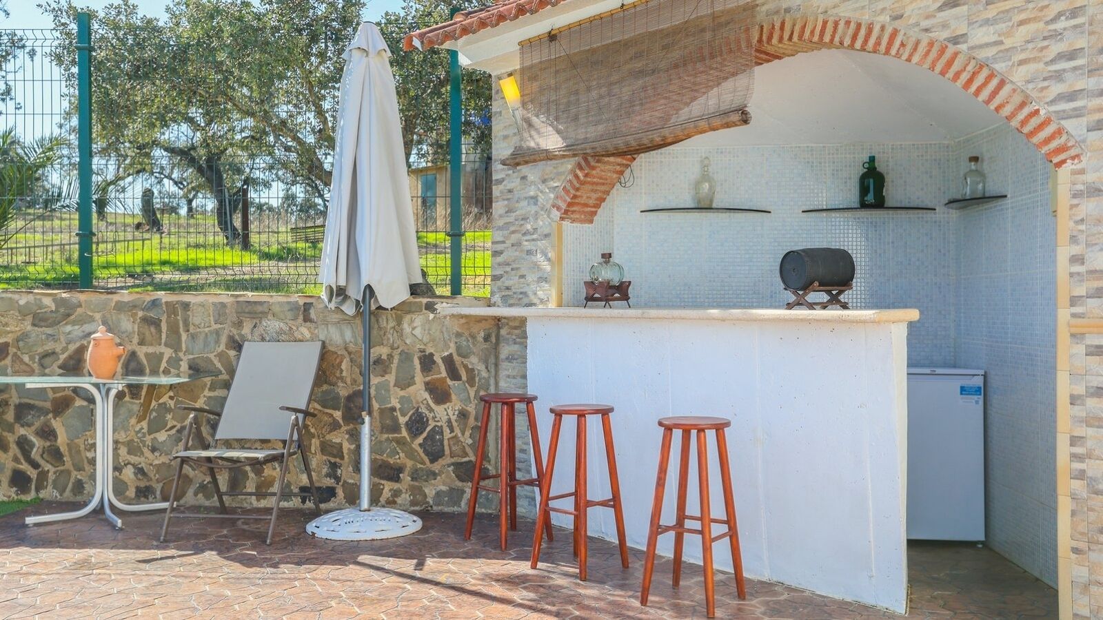 Un área de bar al aire libre con paredes de piedra, un salpicadero de baldosas blancas y una parrilla. Hay tres taburetes y una mesa con una silla.