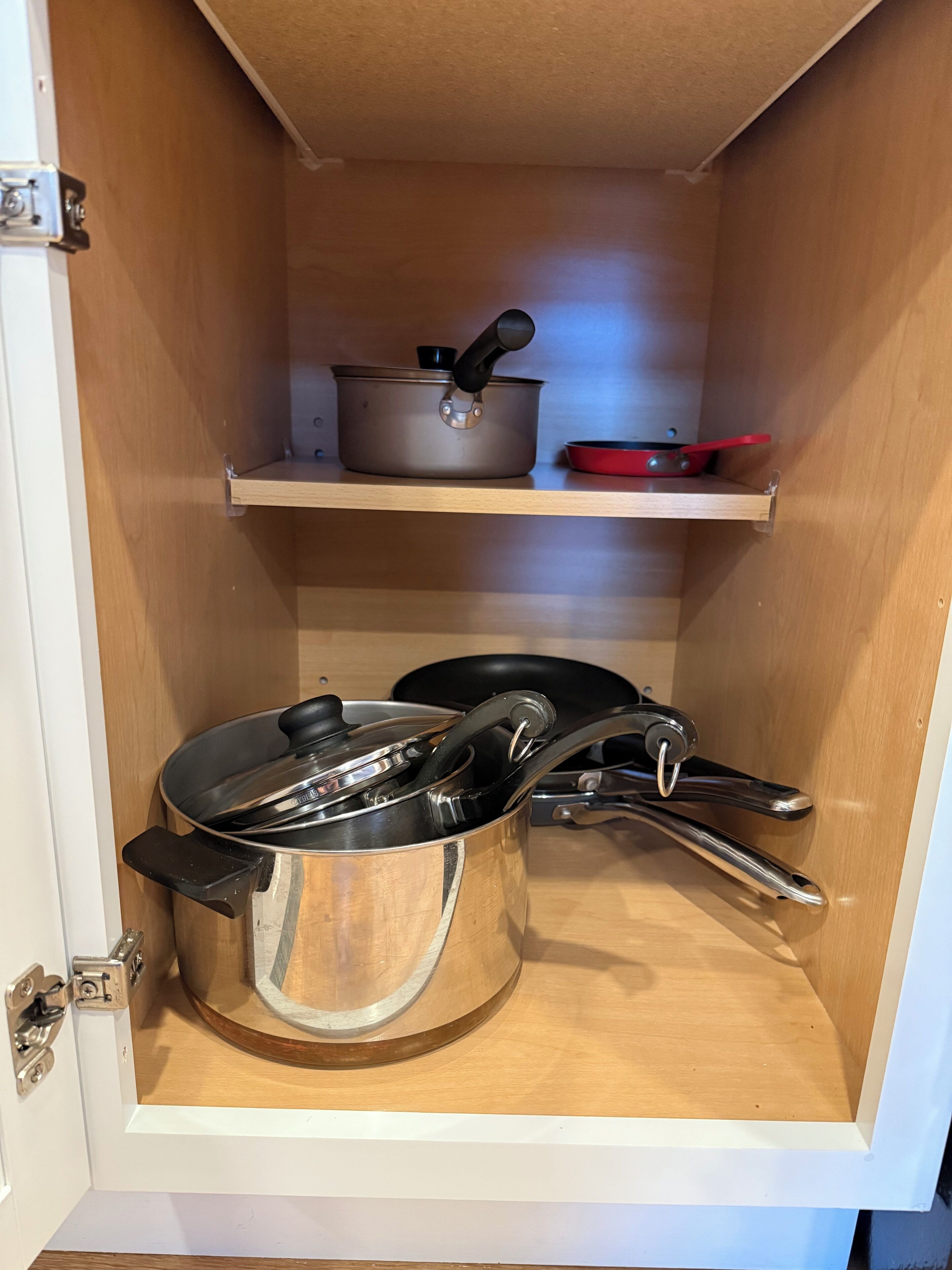 A kitchen cabinet with two stainless steel pots and a red frying pan on a shelf.