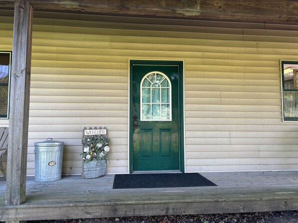 A green door with a window, a welcome sign, a metal trash can, and a flower pot on a porch. - Charming 1-bedroom apartment in Greene for up to 4 guests, family & dog friendly (Greene)