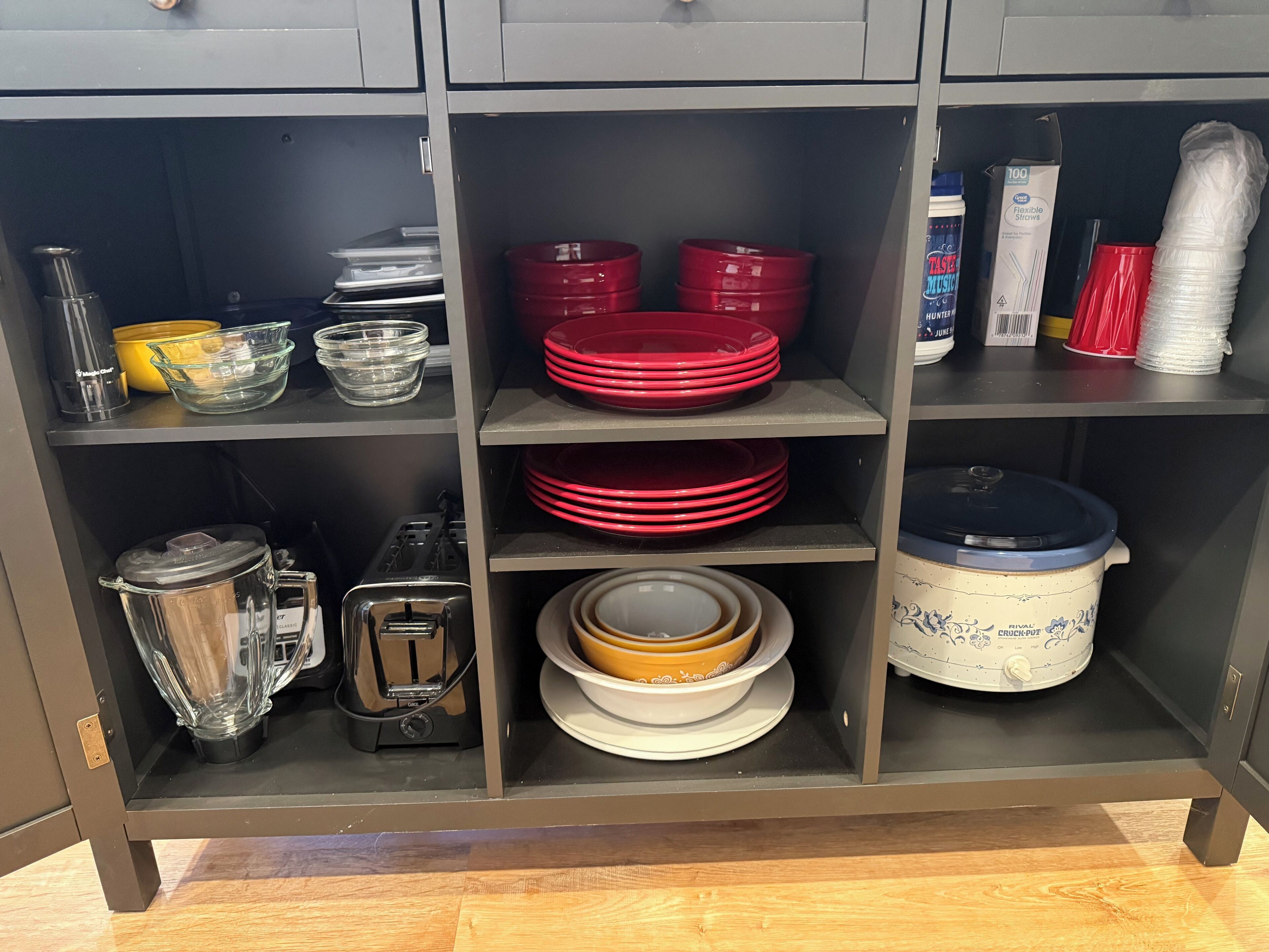 Ein Küchenschrank mit Regalen, voll mit verschiedenem Küchenzubehör, darunter rote Teller, Schüsseln, ein Mixer, ein Toaster und ein Slow Cooker.