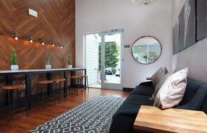 Un intérieur moderne avec un parquet en chevrons, un coin bar avec des tabourets, un canapé noir avec des coussins, un miroir rond et un tapis à motifs.