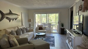 A spacious living room with a large sofa, a coffee table, and a television. There is a balcony with a view of greenery. - Stunning 2 bedroom, 2 balcony condo on golf course with tennis and pickleball (Naples)