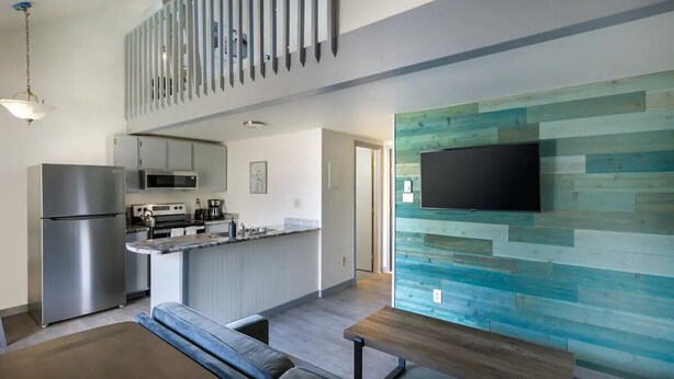 A modern kitchen with stainless steel appliances, a dining area with a wooden table and bench, and a living room with a large TV mounted on a wooden wall.