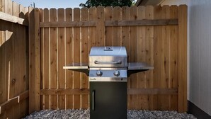 A Char-Broil gas grill with two burners and a side shelf, placed on gravel in front of a wooden fence.