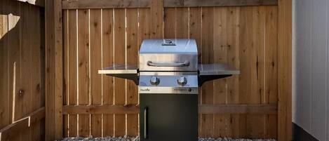 Una parrilla de gas Char-Broil con dos quemadores y una repisa lateral, ubicada sobre grava frente a una cerca de madera.
