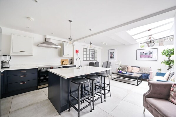 A modern kitchen with a central island, bar stools, a dining area with a sofa, and a large window providing natural light.