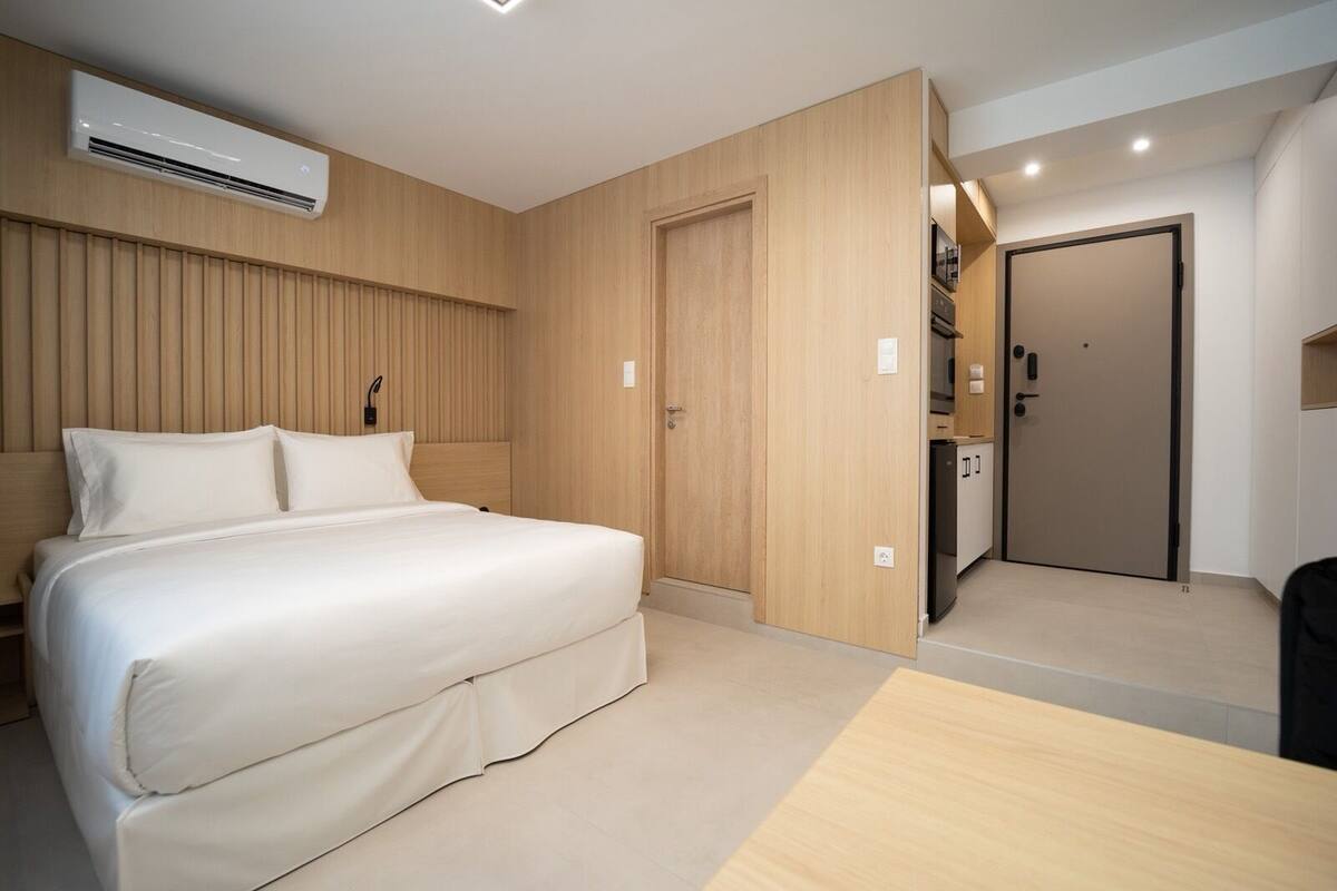 Una habitación de hotel moderna con un panel de madera en la pared, una cama con ropa blanca, un equipo de aire acondicionado integrado y una pequeña zona de kitchenette.