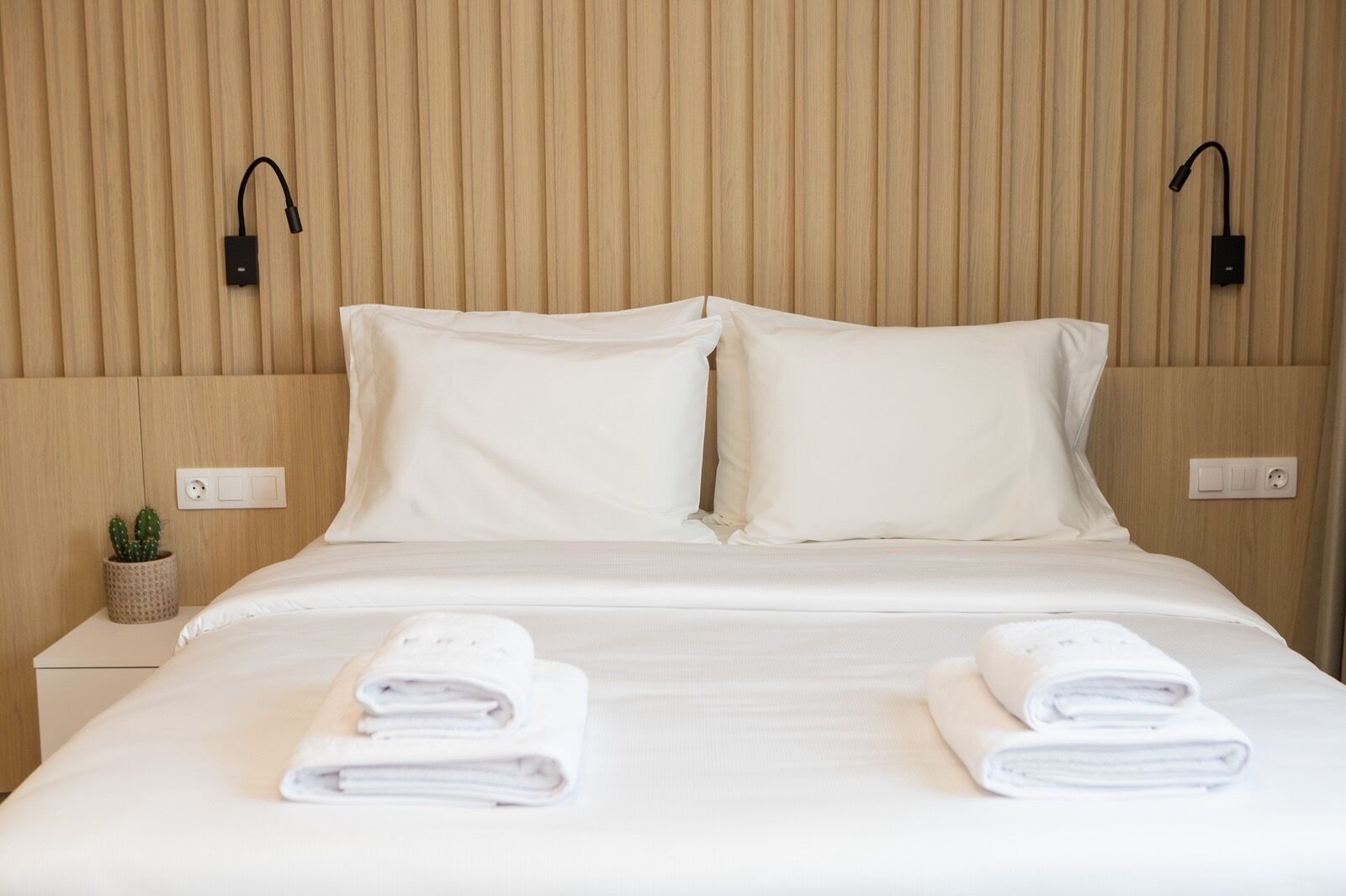 Un lit bien fait, avec du linge de lit blanc et deux oreillers, une petite plante en pot sur la table de chevet et deux lampes de lecture fixées au mur.