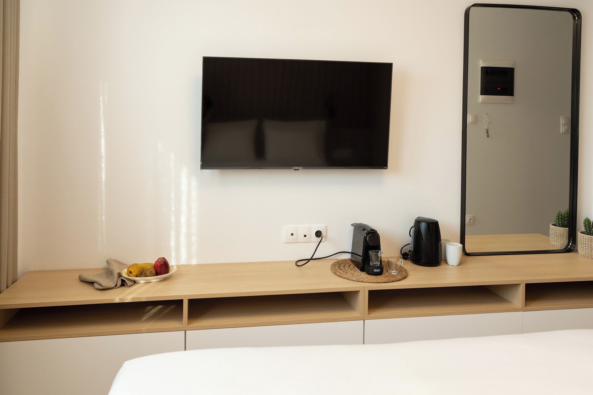 A modern hotel room with a flat-screen TV mounted on the wall, a bedside table with a fruit bowl, a coffee maker, and a mirror.