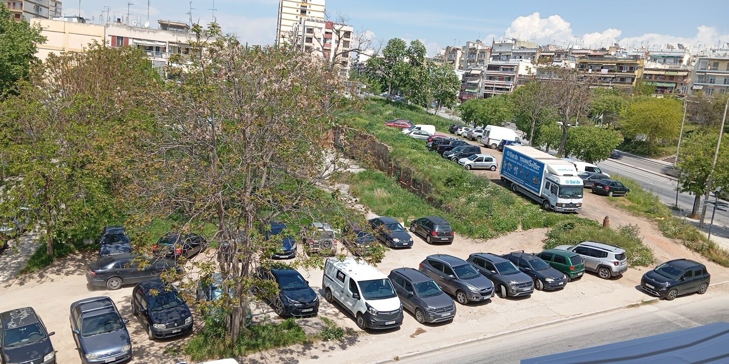 A parking lot with multiple cars and a white van, surrounded by trees and buildings in the background.