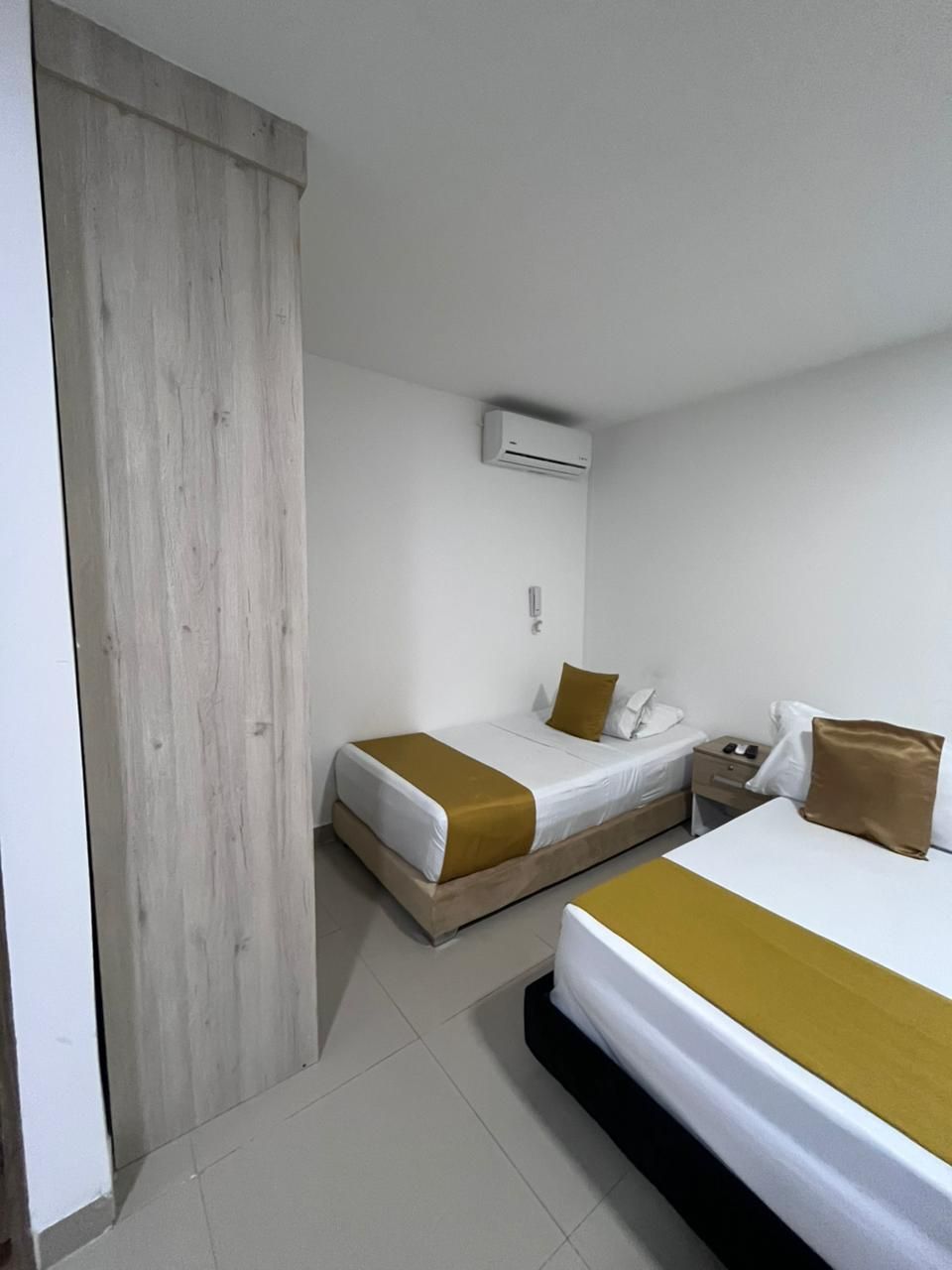 Habitació d'hotel amb dos llits, una porta de fusta, un aparell d'aire acondicionat blanc i una tauleta de nit amb un telèfon.