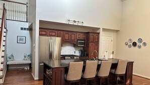 A kitchen with a central island, wooden cabinets, a stainless steel refrigerator, and a dining area with bar stools. - Enjoy Derby City in this amazing river bungalow! (Louisville)