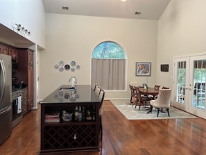 A kitchen with a central island, dining area with chairs, a large window with a view, and a door with glass panels.