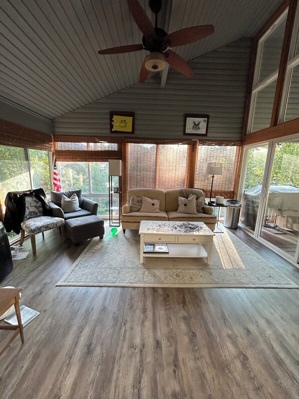A spacious living room with a ceiling fan, a sofa set, a coffee table, and large windows.