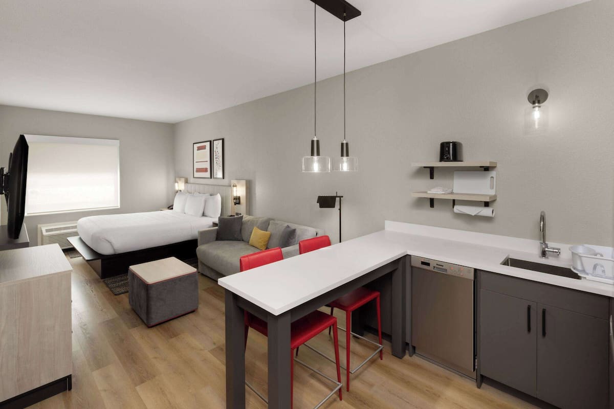 A modern hotel room with a bed, a sofa, a dining area, and a kitchen.