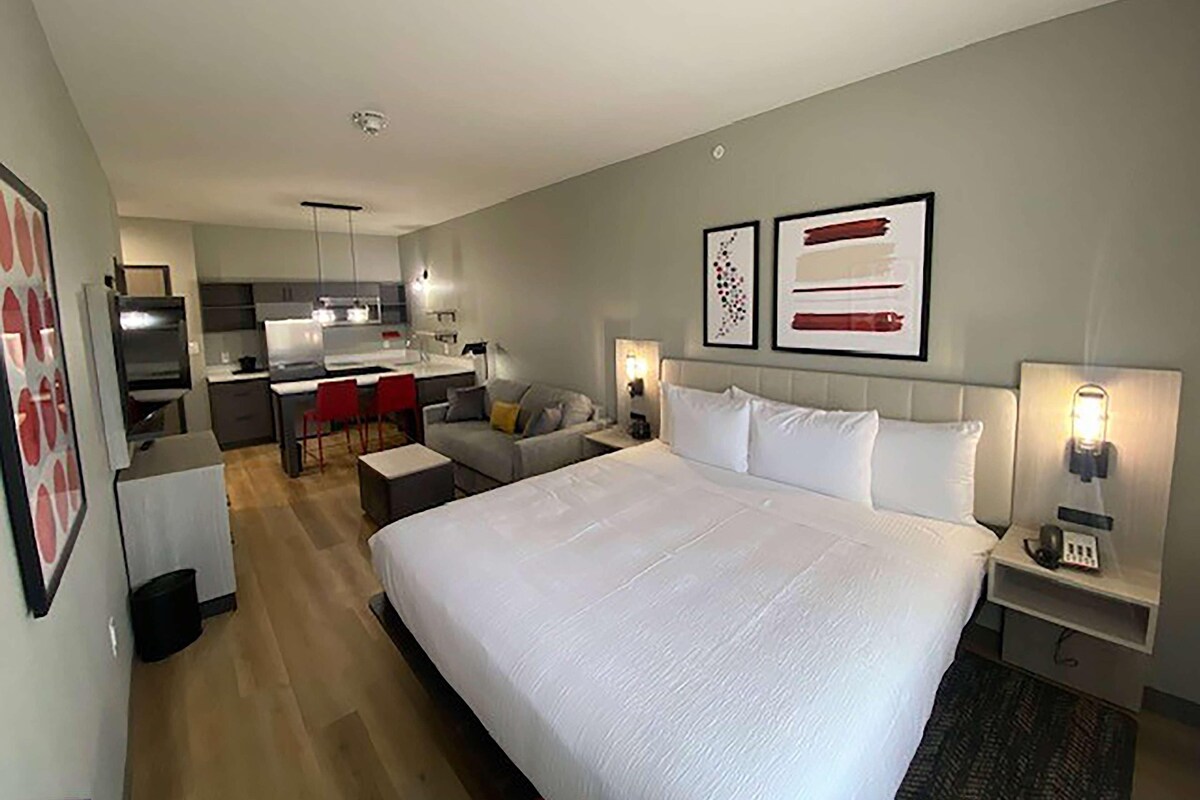A modern hotel room with a large bed, a kitchenette, a dining area, and a sitting area.