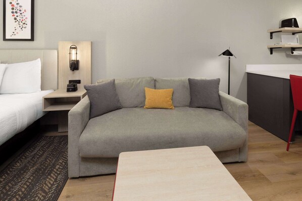 A modern hotel room with a grey sofa, a small table, a bedside table with a lamp, and a wall-mounted shelf. - Hawthorn Extended Stay by Wyndham DeSoto (DeSoto)