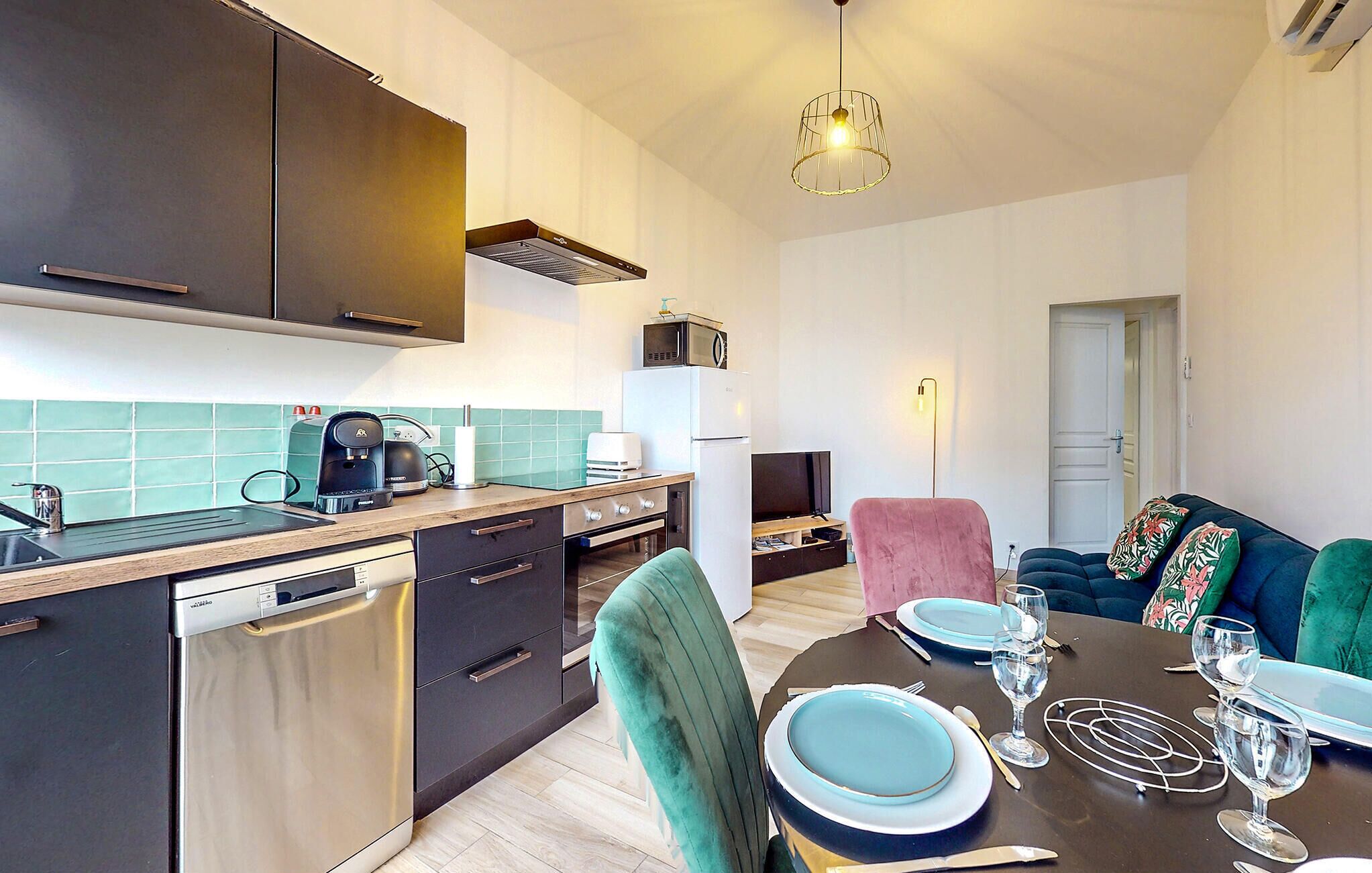 Een moderne keuken met een vaatwasser, magnetron en koffiezetapparaat. Er is een eethoek met een tafel gedekt voor twee personen, en een bank op de achtergrond.