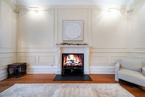 Een woonkamer met een open haard, een ingelijst kunstwerk erboven, een bank en een houten bijzettafeltje.