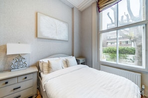 Quarto com cama, mesa de cabeceira, abajur e janela com vista para edifícios e árvores.