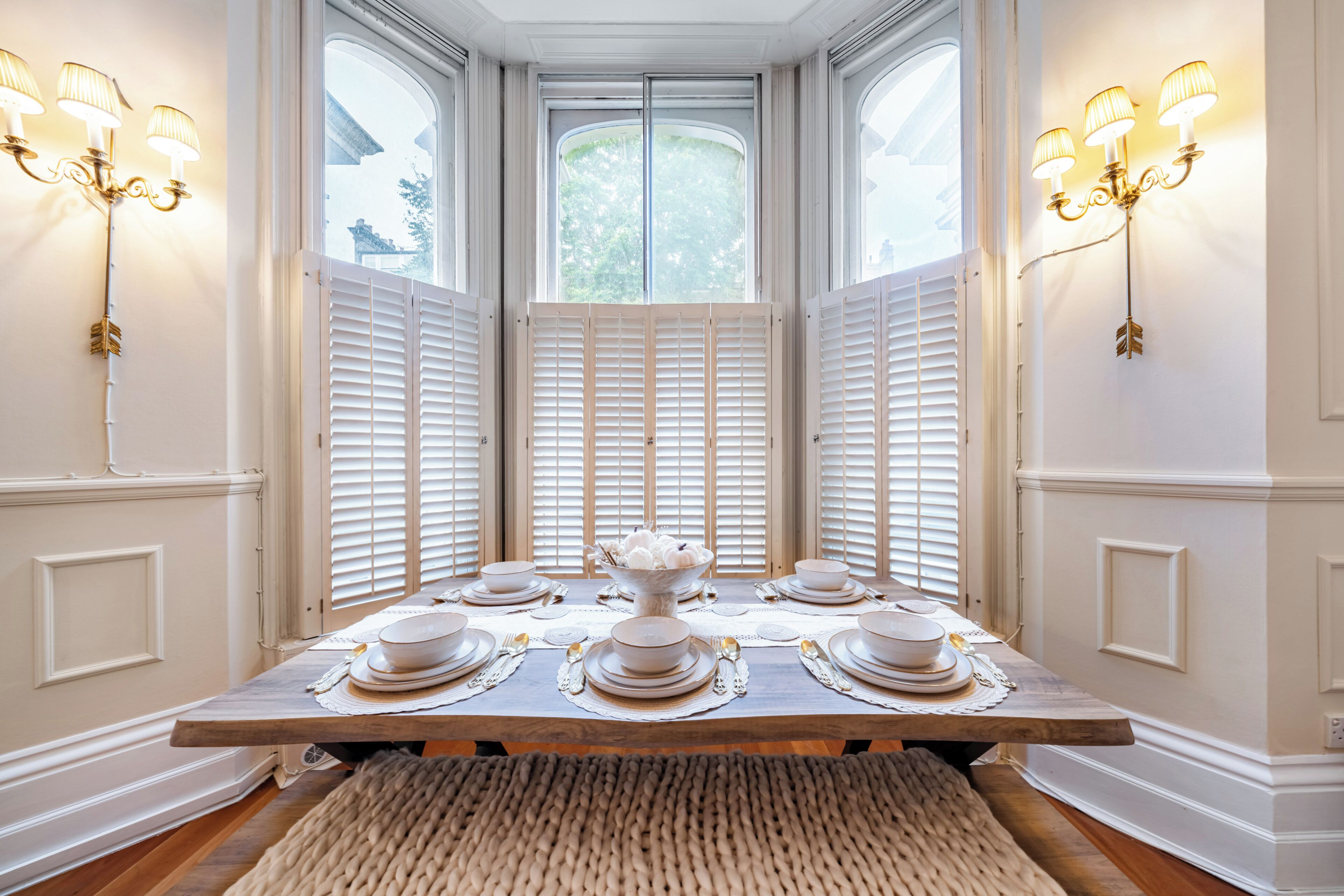 A dining table set for six with white plates, bowls, and silverware, flanked by two wall-mounted light fixtures.