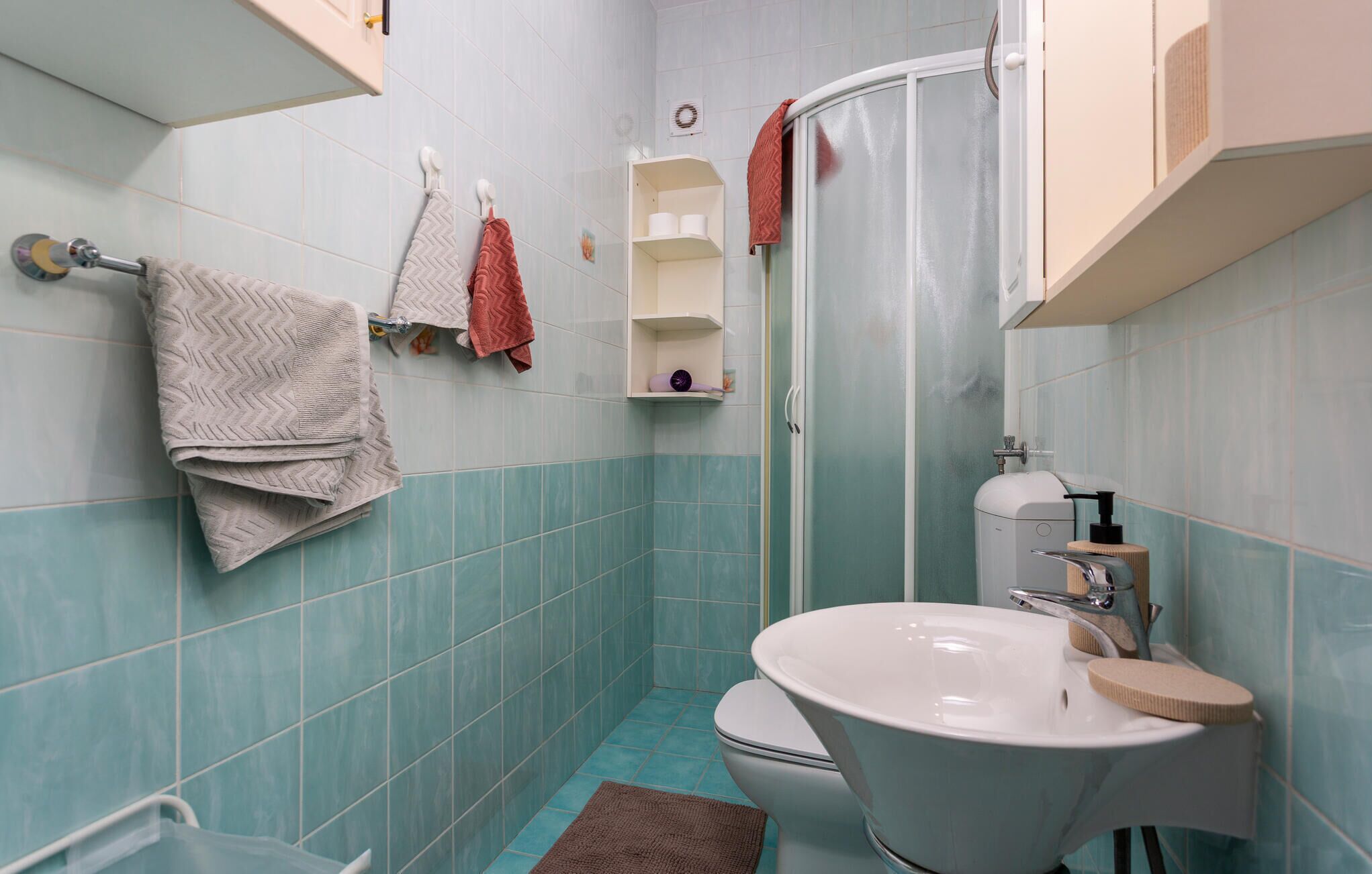 Un baño con inodoro, lavabo y ducha. Hay toallas colgadas en la pared y un estante con artículos de aseo.