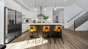 A modern kitchen with a stainless steel refrigerator, wooden bar stools, pendant lights, and a marble countertop.