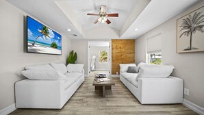 A modern living room with two white sofas, a wooden accent wall, a ceiling fan, and a large TV displaying a beach scene.