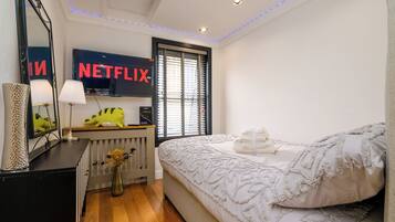 A bedroom with a bed, a dresser, a mirror, a radiator, a wall-mounted TV displaying the Netflix logo, and a window with blinds.