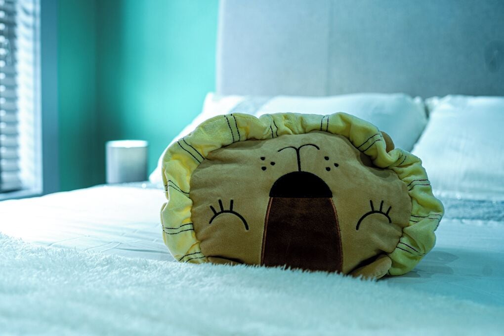 A plush toy resembling a bear's face on a bed with a white blanket and a cup in the background.